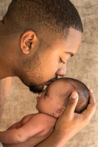 Nouveau né dans les bras de son père avec un bisous sur le front du bébé -photographe de naissance- krystyne ramon