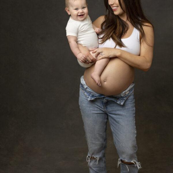 future maman avec son jean ouvert sur le ventre tenant un autre bébé dans ses bras -photographe de naissance