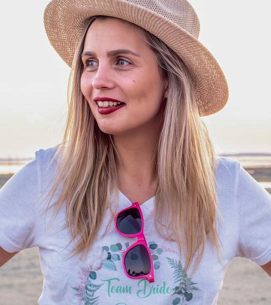 séance EVJF portrait d'une jeune fille sur la plage un chapeau de paille sur la tête et de lunettes rose accrochées sur son tee shirt krystyne ramon - home studio photos