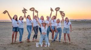 séance EVJF groupe de dix jeunes filles sue la plage un bras en l"air avec un chapeau dans la main et les lettre EVJF posées sur le sable