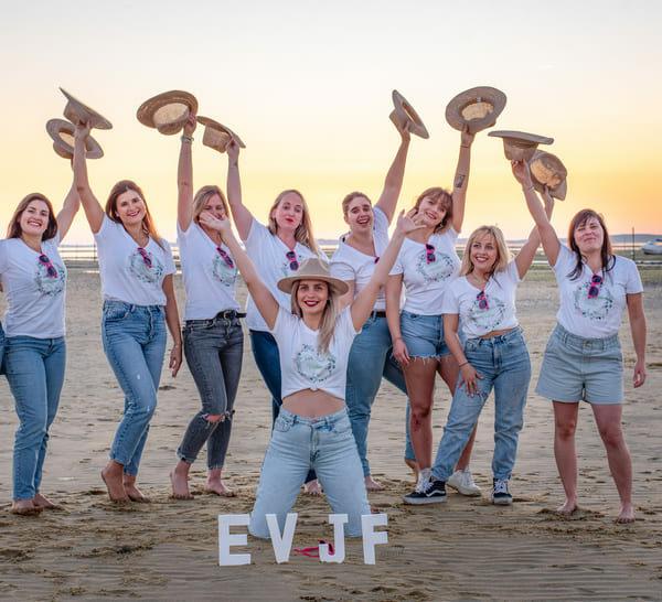 séance EVJF groupe de dix jeunes filles sue la plage un bras en l"air avec un chapeau dans la main et les lettre EVJF posées sur le sable krystyne ramon - home studio photos