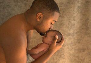 Nouveau né dans les bras de son père avec un bisous sur le front du bébé -photographe de naissance- krystyne ramon